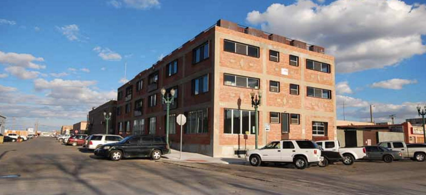 This image shows a large, three-story brick building with many dark-framed windows situated on a street corner under a blue sky filled with scattered clouds. A row of parked vehicles, mostly SUVs and pickup trucks, lines the curb in front of the building, with additional cars further down the street. The area appears to be part of an urban or industrial district, as evidenced by the plain, functional architecture and adjacent low-rise buildings. Green street lamps are positioned along the sidewalk, and a stop sign is visible at the corner. The scene is brightly lit by afternoon sunlight, casting distinct shadows on the road and buildings.